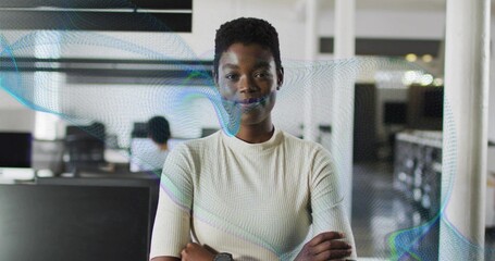 Standing woman in beige top crossing arms facing camera in open-plan office, monitors overlay watch