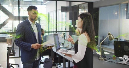 Discussing pair wearing dark blazer and light blouse, standing in office holding laptop and papers
