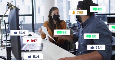 Discussing woman masked in cardigan and man in shirt at laptop in office with papers, alerts