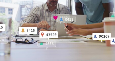 Gesturing mature man in checkered shirt pointing at laptop in office, with docs, pen, social icons