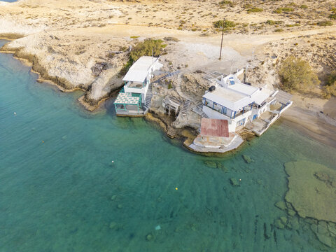 Aerial view of buildings perched on the edge where the rugged coastline meets the crystal-clear turquoise waters, Paralia Mitikas, Milos, Greece.