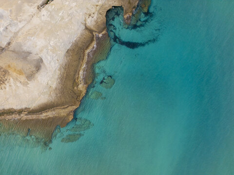 Aerial view of a rugged coastline meeting the turquoise sea, where the light sand contrasts with the clear, vibrant water, Paralia Mitikas, Milos, Greece.