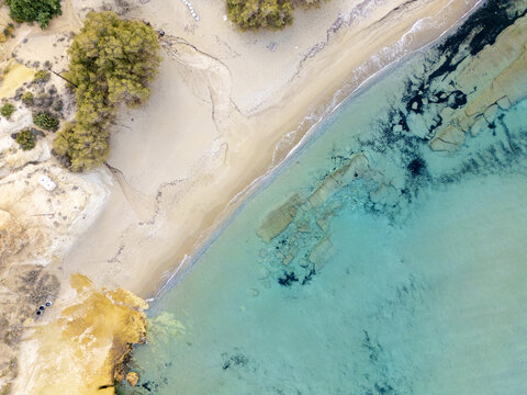 Aerial view of azure waters meeting the soft sandy shore near rocky outcrops and sparse vegetation, Paralia Mitikas, Milos, Greece.