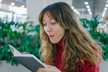 Young woman with long curly hair and a red sweater feeling surprised and amazed while reading a book, discovering something new, and expanding her knowledge at a library or public space