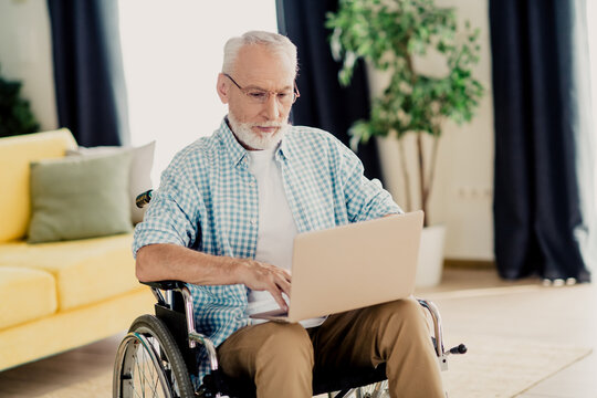 Elderly man in wheelchair works on a laptop at home, showcasing inclusion and disability awareness in a cozy living room setting - Powered by Adobe