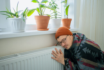 Man in warm clothes huddled by a radiator in a chilly apartment, wrapped in a blanket and beanie, illustrating winter heating struggles, high energy bills and financial stress