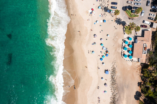 Aerial view of turquoise waves crashing onto the sandy shore alongside colorful umbrellas and a parking lot filled with cars, Laguna Beach, California, United States.