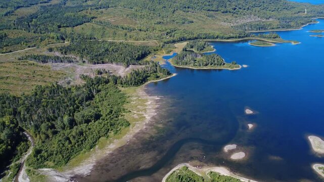 Aerial view of a lake bordered by dense forests, logging camps, and a forest road running along a winding shoreline dotted with islands and small coves. Lac-a-la-Croix, La Mitis, Quebec, Canada, 2025.