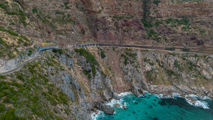 Aerial view of the dramatic Chapmans Peak Drive clinging to the rugged coastline where turquoise waters meet towering cliffs, Cape Town, Western Cape, South Africa.