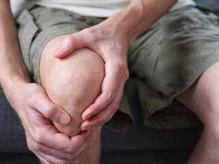 A middle-aged man sitting on a couch, holding his knee and leg with both hands in pain. Close up.
