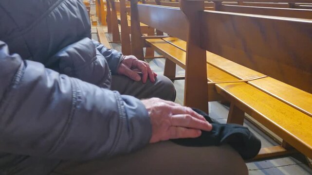 A man sits quietly on a wooden pew of church holding a black cap in his hands