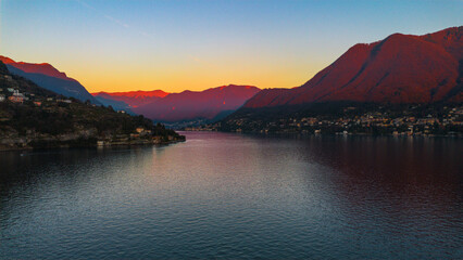 Aerial view of the tranquil lake mirroring the fiery sunset hues painting the distant mountains, a serene vista of nature's artistry, Como, Lombardy, Italy.