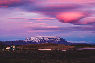 Aerial view of stark black earth meets the tranquil lake, while the church spire points to the vibrant hues of the sunset sky, Reykjahlid, North Iceland, Iceland.