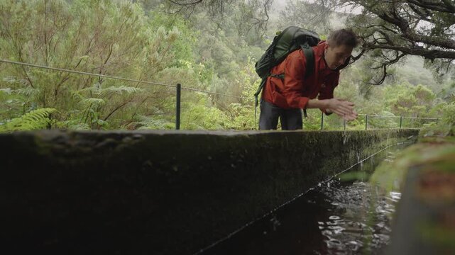 A close, natural shot of a male hiker washing his hands and face in a flowing levada along the PR6 Levada das 25 Fontes trail in Madeira. Clear mountain water runs through the narrow channel