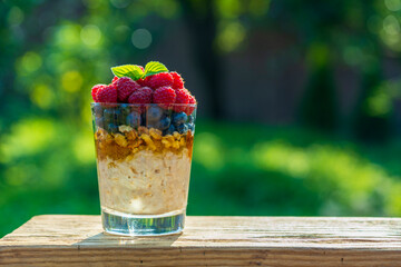 Summer breakfast layered dessert in a glass of oatmeal, blueberries, raspberry, walnut, olive oil and honey on nature background, closeup. Healthy diet with fresh berries on wooden table
