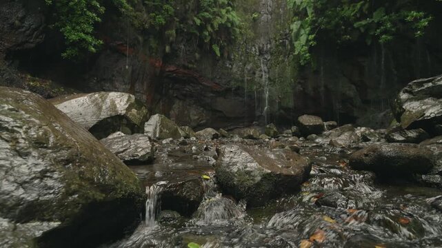 A static, low-angle shot at the 25 Fontes waterfall in Madeira. Wet rocks and flowing water fill the foreground, while multiple thin streams cascade down the cliff in the background.
