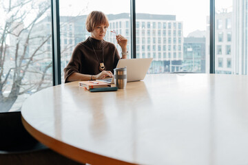 White woman aged 40 reviewing documents on laptop at conference table by window