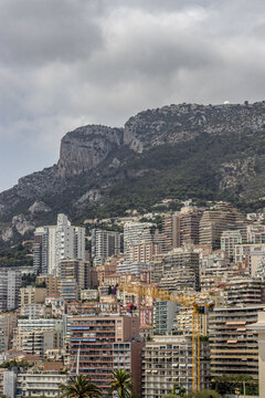 View of buildings rise in a dense urban landscape against a backdrop of rugged, mountainous terrain under a cloudy sky, Monaco, Monaco.