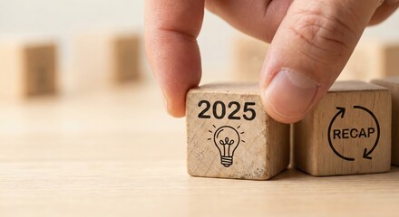 Close-up of a white adult hand turning wooden blocks on a light desk showing 2025 recap text and lightbulb icon, representing annual reflection and creative planning in a bright indoor setting.