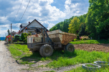 Obraz premium Heavy dump truck parked on grass near a rural road and freshly worked field under a bright sky. Countryside construction and agriculture concept showing machinery, transport, and seasonal outdoor work