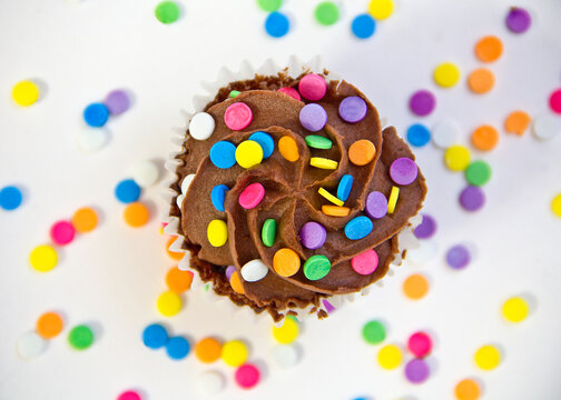 Close-up overhead view of a chocolate cupcake with buttercream frosting and multi coloured sprinkles on a table with more sprinkles