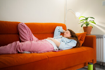 Caucasian woman resting on a cozy orange couch feeling unwell with toxicosis symptoms, experiencing weakness and nausea in a domestic setting, wearing comfortable pajamas at home