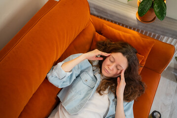 Young woman suffering from a headache or migraine, lying on an orange sofa, pressing her temples, eyes closed in discomfort, reflecting symptoms of illness, stress, and chronic pain at home
