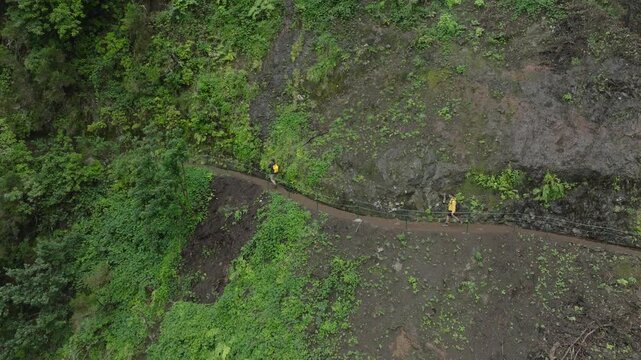 A drone shot of the PR9 Levada trail carved into a steep vertical cliff on Madeira Island. Dense levada vegetation covers the rock wall, while the narrow water channel and walking path are are clearly