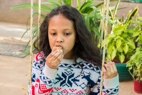 Little girl enjoying a traditional Indian sweet laddu while sitting on a garden swing. joyful childhood moment in an outdoor setting, family lifestyle, traditional food, cultural taste