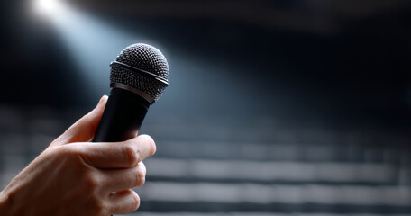 Close-up of hand holding microphone under spotlight with blurred empty auditorium seats in background