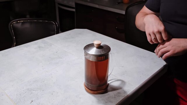 Male hands pressing a plunger of a French press with a dark tea beverage brewing in a glass carafe on kitchen table on dark background. Pressed sediment falls. Evening ritual. High quality 4k footage.