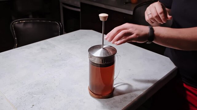 Male hands lift a plunger of a French press with a dark tea beverage brewing in a glass carafe on kitchen table on dark background. Pressing sediment falls. Evening ritual. High quality 4k footage.