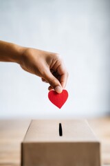Hand placing a red heart symbol into a donation box, representing generosity and kindness, with a soft blurred background enhancing the emotional connection of giving
