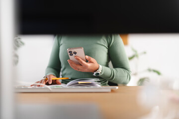 A person uses a smartphone while holding a pencil over an open notebook at a desk, suggesting multitasking and productivity in a modern workspace.
