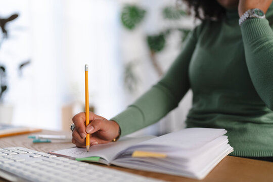 A person is writing in a notebook with a pencil at a desk with a keyboard and a plant in the background.