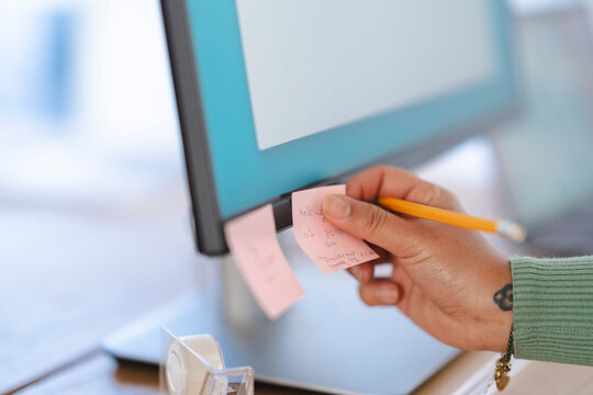 A person's hand holds a pencil and a pink sticky note with writing on it, placing it on a computer monitor.