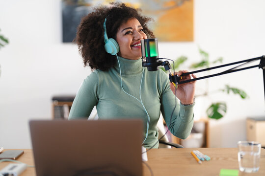 A woman with headphones speaks into a microphone while working on a laptop, creating content for her broadcast.