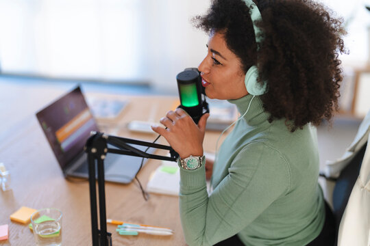 A woman with headphones speaks into a microphone while recording a podcast. A laptop is open on the desk beside her.