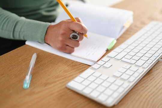 A person's hand, adorned with a ring, writes in a notebook with a pencil next to a keyboard on a wooden desk.