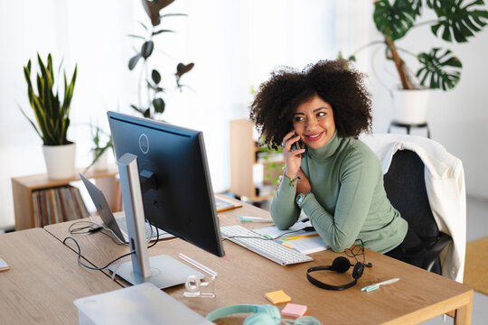 A woman with curly hair smiles while talking on her phone at a desk with a computer and plants.