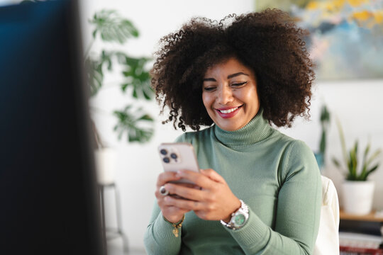 A smiling woman with curly hair looks at her phone while sitting in a bright, modern office space with plants in the background.