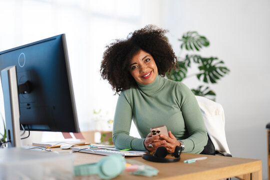 A smiling woman with curly hair sits at her desk, holding a smartphone. She is in a bright office with a computer and plants in the background.