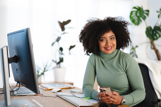A smiling woman with curly hair sits at her desk, holding a smartphone. She is in a bright office with plants in the background.