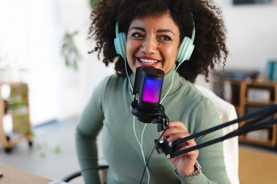A woman with curly hair wears headphones and speaks into a microphone with colorful lights, recording audio for a podcast or broadcast.