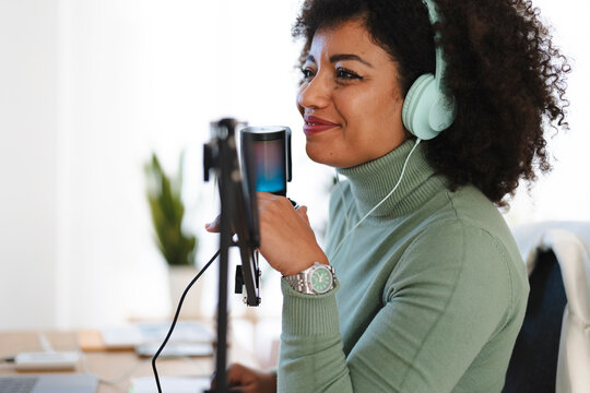 A woman with curly hair wears headphones and speaks into a microphone, recording audio for a podcast or broadcast.