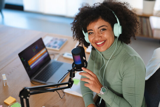 A woman with curly hair wears headphones and smiles while speaking into a microphone at a desk with a laptop.