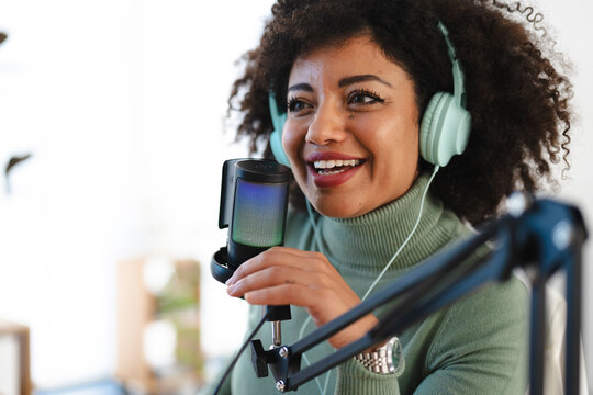A woman with curly hair wears headphones and speaks into a microphone, recording audio for a podcast or broadcast.
