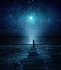 Fototapeta premium Girl facing the ocean under a luminous star in a deep night sky. Mystical, spiritual, cosmic atmosphere with water reflection and celestial light. Elements of this image furnished by NASA