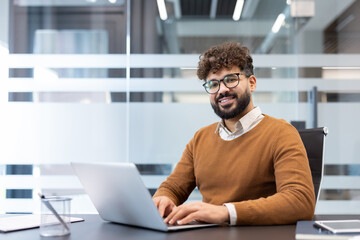 Obraz premium Young man with beard and glasses smiling at camera, sitting at a desk and using a laptop in a contemporary office environment, embodying professionalism and success