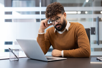 Young bearded businessman in glasses concentrating on his laptop at a modern office desk, thoughtfully analyzing data and planning solutions for a corporate project or strategy